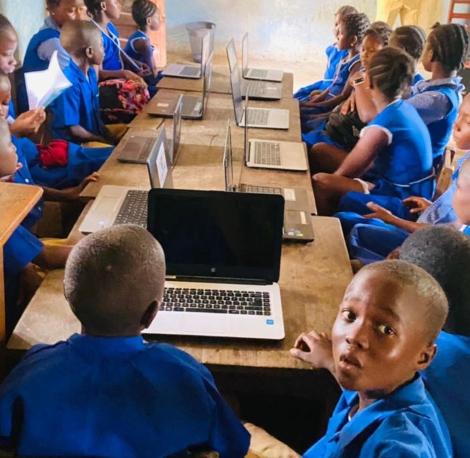 Pupils in blue school uniforms seated along rows of desks with laptops in a classroom